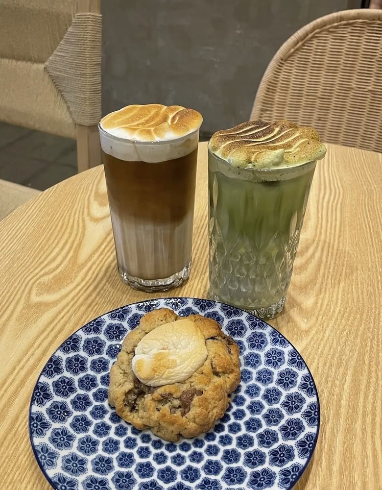 Deux cloud lattes, café et matcha, coiffés de meringue toastée, avec un cookie sur une assiette fleurie bleue.