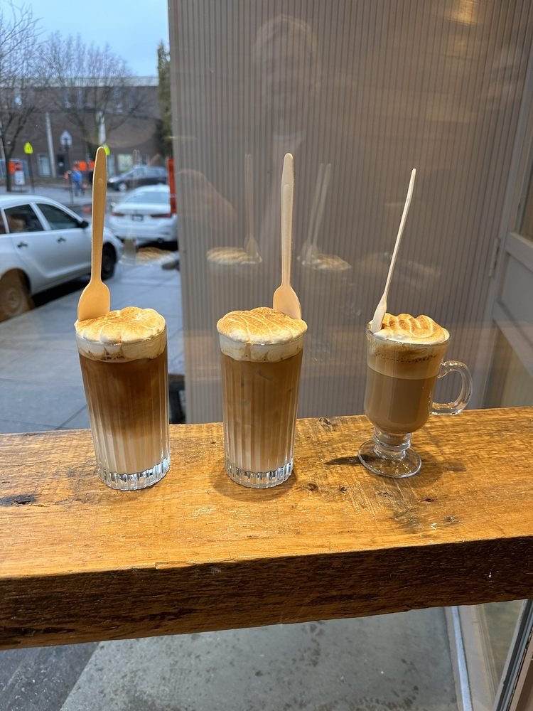 Trois cloud lattes alignés sur le rebord de fenêtre, chacun surmonté d'une cuillère en bois plantée dans la meringue.