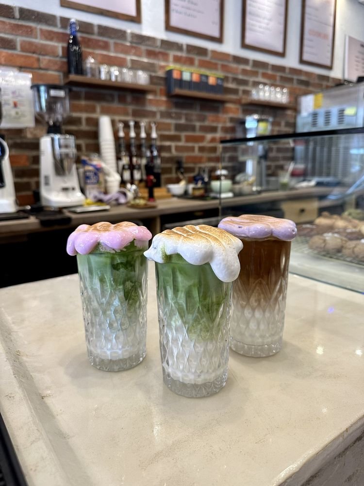 Trois matchas coiffés de meringue colorée alignés sur le comptoir, devant le mur de brique.